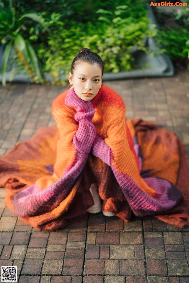 A woman sitting on a brick walkway wrapped in a blanket.